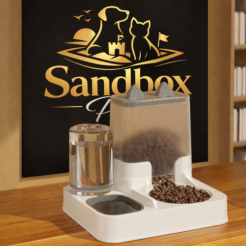 A white automatic pet feeder and water dispenser set on a wooden table, with a clear water glass and a transparent food hopper filled with dog food. A black wall behind it displays a large gold 'Sandbox' logo.