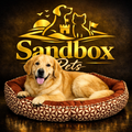 A golden retriever dog lies in a brown patterned pet bed with a brown cushioned interior, set against a dark textured background with a large gold 'Sandbox Pets' logo and silhouettes of a dog, cat, and castle.