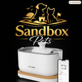 A white pet water fountain with a wooden base and a clear lid, shown with a smartphone app screen and a large gold 'Sandbox Pets' logo on a black background.