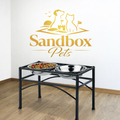 A black metal elevated pet feeder stand with two stainless steel bowls, one filled with food, shown in a room setting on a wood floor against a white wall with a gold 'Sandbox Pets' wall decal above.